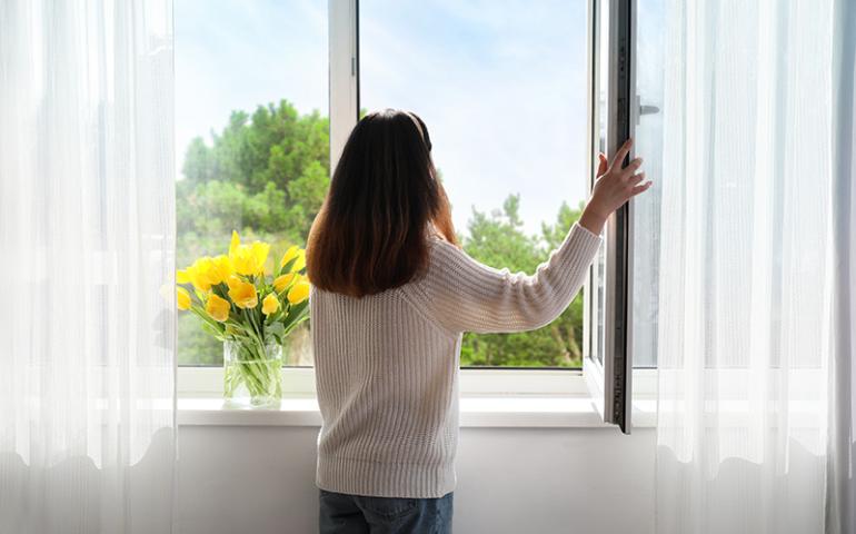 Woman opening a window with lots of trees and greenery outside.